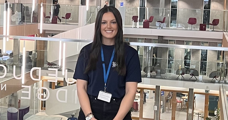Trainee Manager Holly Henderson in front of a glass awards cabinet promoting Apprenticeship Week