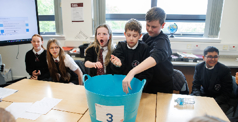 Students at Trinity High School gather around a large blue tub during a classroom challenge; one student reaches into the tub while others look on with excitement and surprise.