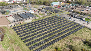 An aerial view of a solar farm with panels mounted on top of grass. In the background of the image is a waste water treatment works.