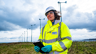 Scottish Water employee in PPE holding a tablet in front of a wind farm
