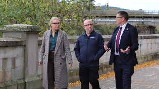 A woman in a long coat is pictured left of the image, with two gentlemen in the centre and right of the picture. They're walking down a pavement with stone wall and trees in the background.