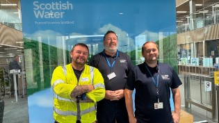 Three employees standing in front of a Scottish Water banner inside an office building. The person on the left is wearing a high-visibility vest, and the other two are in casual work attire, all smiling at the camera.