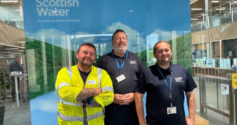 Three employees standing in front of a Scottish Water banner inside an office building. The person on the left is wearing a high-visibility vest, and the other two are in casual work attire, all smiling at the camera.