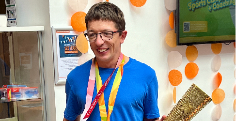 Paralympian Scott Quin smiling while holding a Paralympic torch and wearing his Paralympic medals, with orange and white decorations in the background.